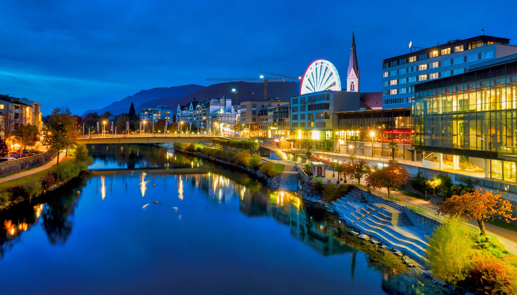 Villach bei Nacht, Panorama von der Drau mit einer Brücke darüber und dem Flussboulevard auf rechter Seite, im Hintergrund ein Kirchturm und ein Riesenrad, alles in farbiger Straßenbeleuchtung erhellt