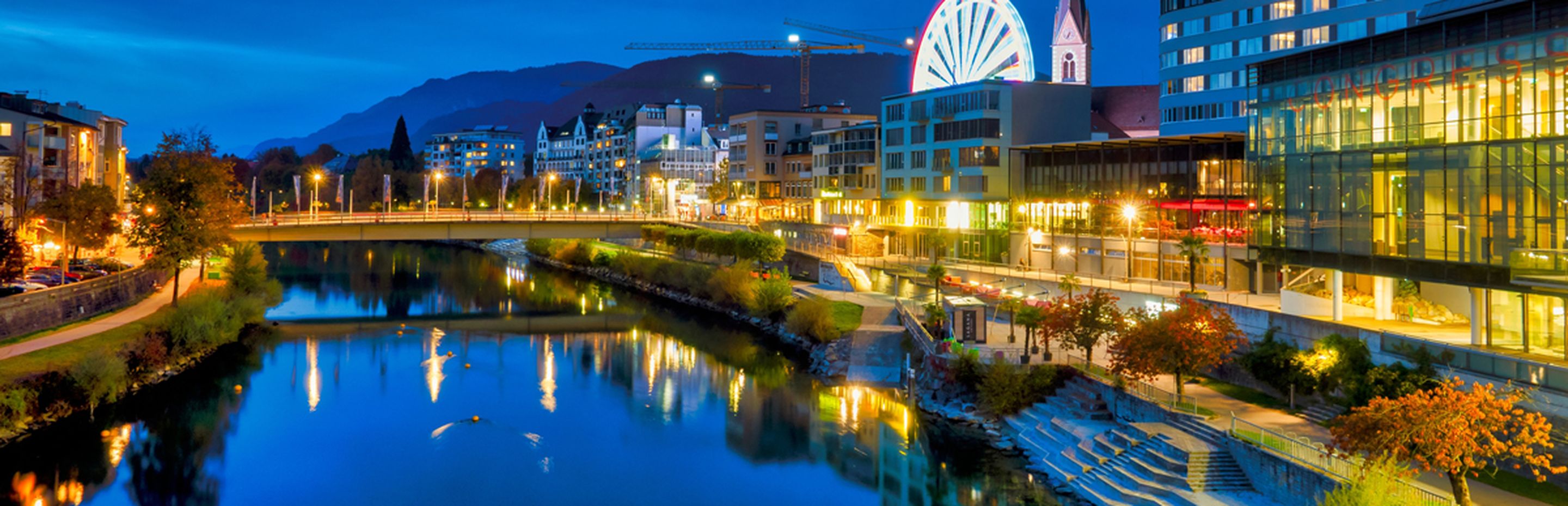Villach bei Nacht, Panorama von der Drau mit einer Brücke darüber und dem Flussboulevard auf rechter Seite, im Hintergrund ein Kirchturm und ein Riesenrad, alles in farbiger Straßenbeleuchtung erhellt - Only Fabrizio / Shutterstock.com