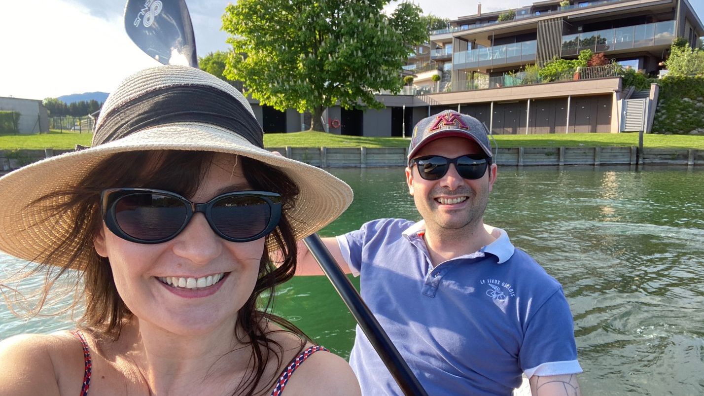 Adelina Shickova und Stéphane Hatterer sitzen in einem Ruderboot und paddeln gerade von Ufer weg, im Hintergrund ein Haus und ein Baum auf grüner Wiese-Adelina Shickova