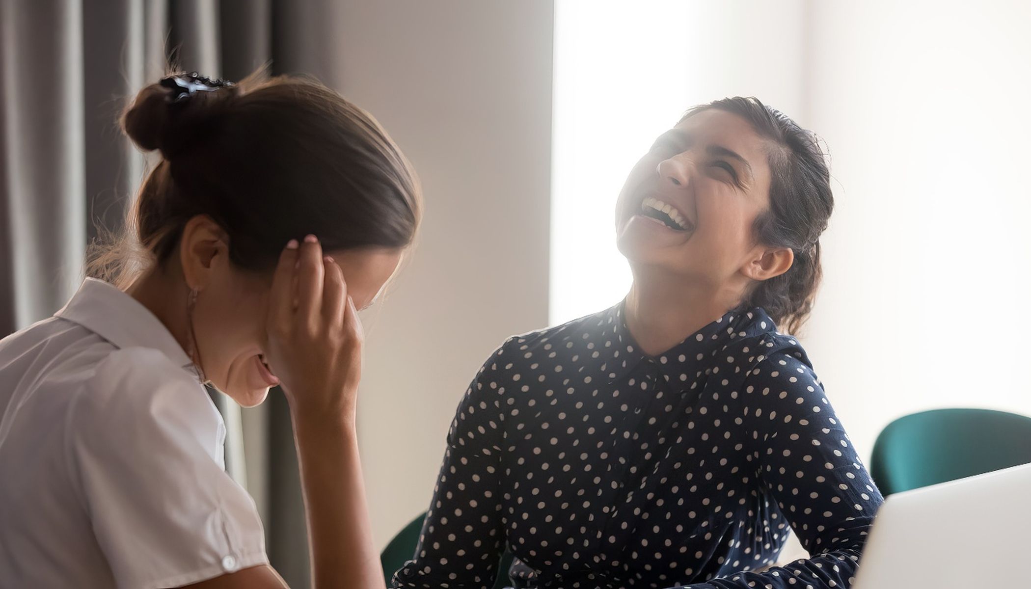 Two women are sitting in the house talking. One is wearing a polka-dot blouse and is laughing openly, the other, in a white shirt, is covering her face with her hand and looks amused. A laptop is partially visible on a table.