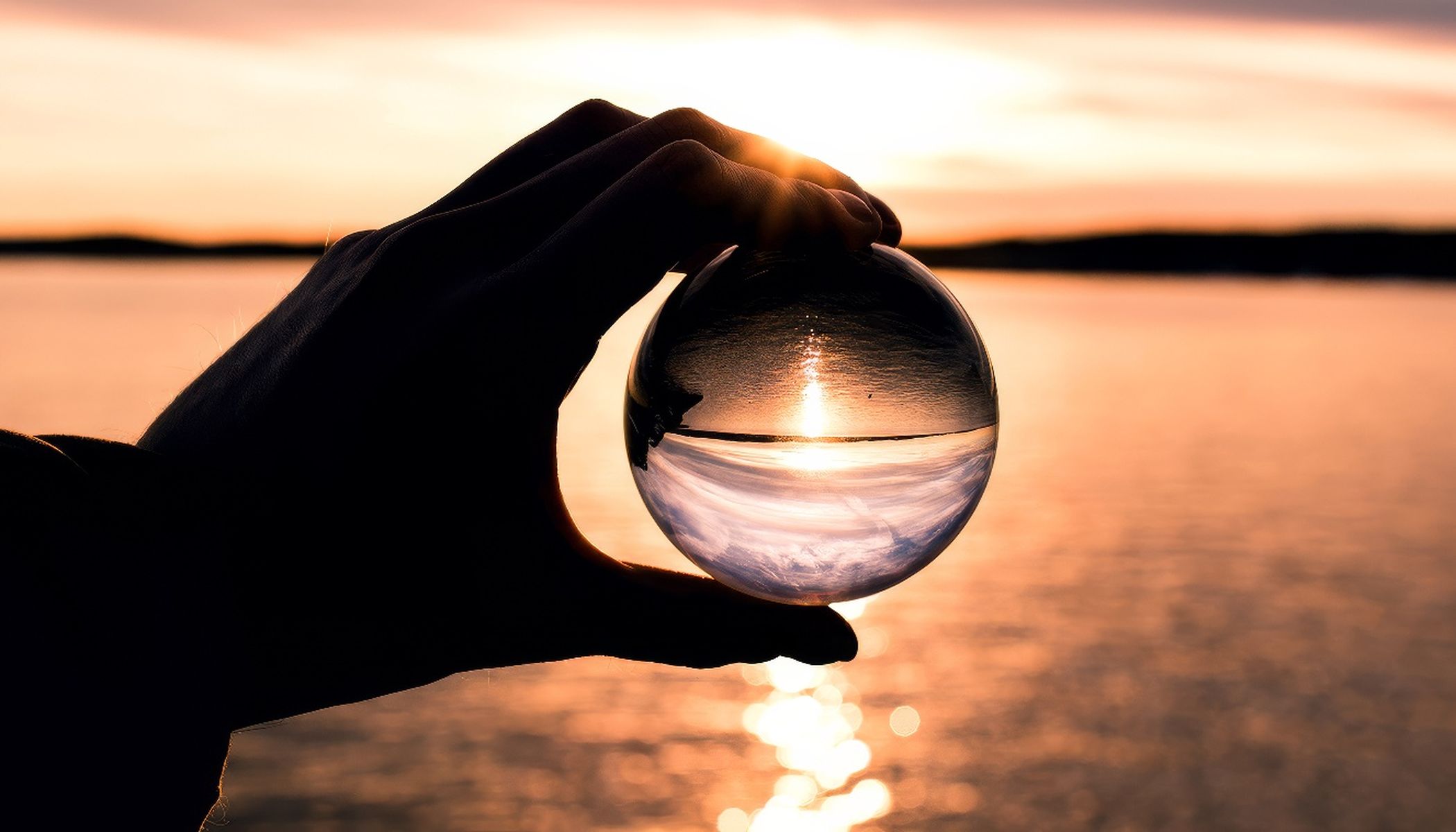 Eine Hand hält eine transparente Kugel, die einen Sonnenuntergang über einem Gewässer reflektiert. Die Sonne steht tief am Horizont, und der Himmel ist in Orange- und Rosatönen gefärbt. Das Wasser darunter glitzert im Spiegelbild der Sonne.