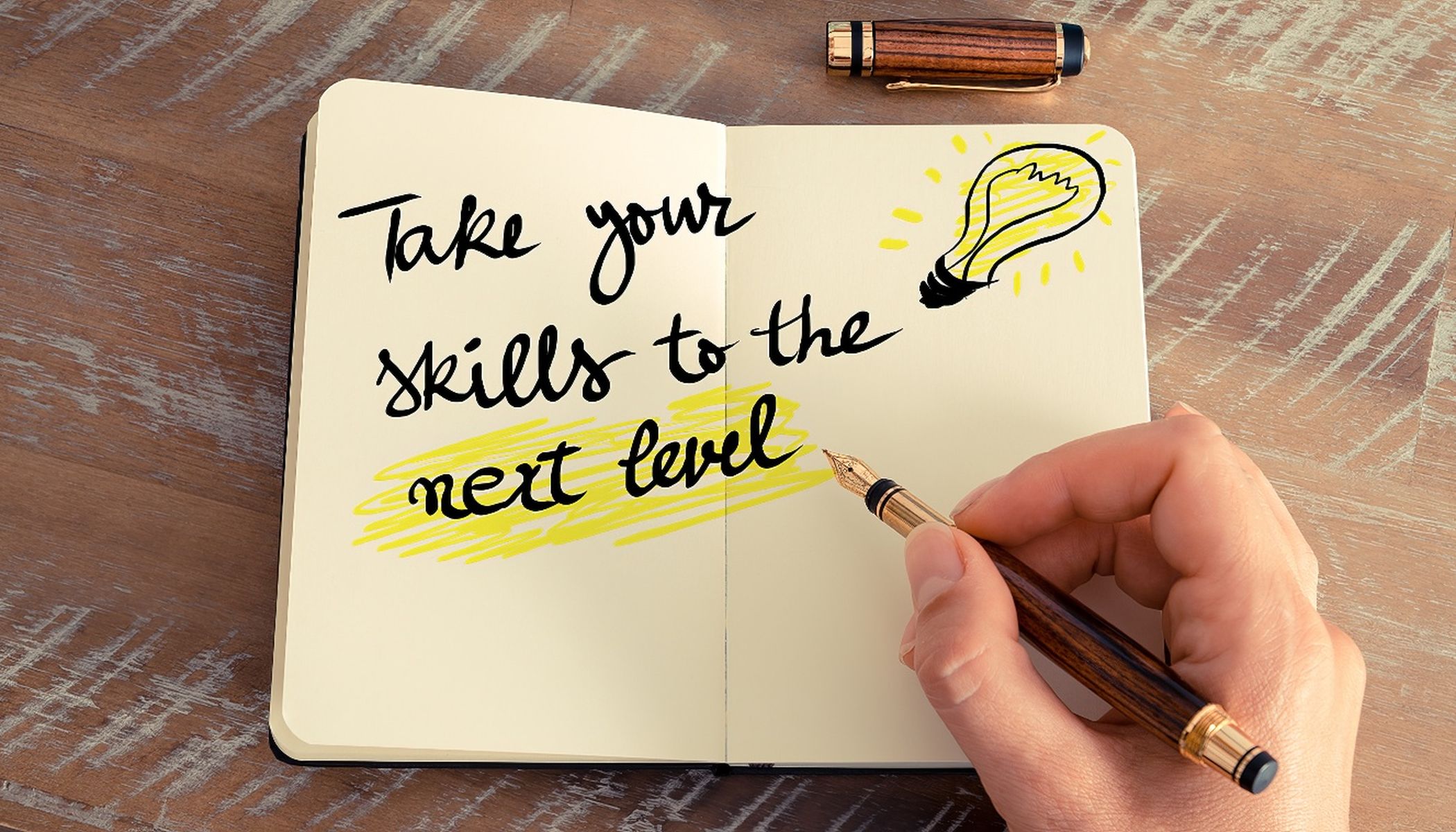 A person writes "Take your skills to the next level" in a notebook with a pen. The text is highlighted in yellow, with a light bulb drawn next to it. The notebook lies on a wooden surface and the pen has a wooden design.