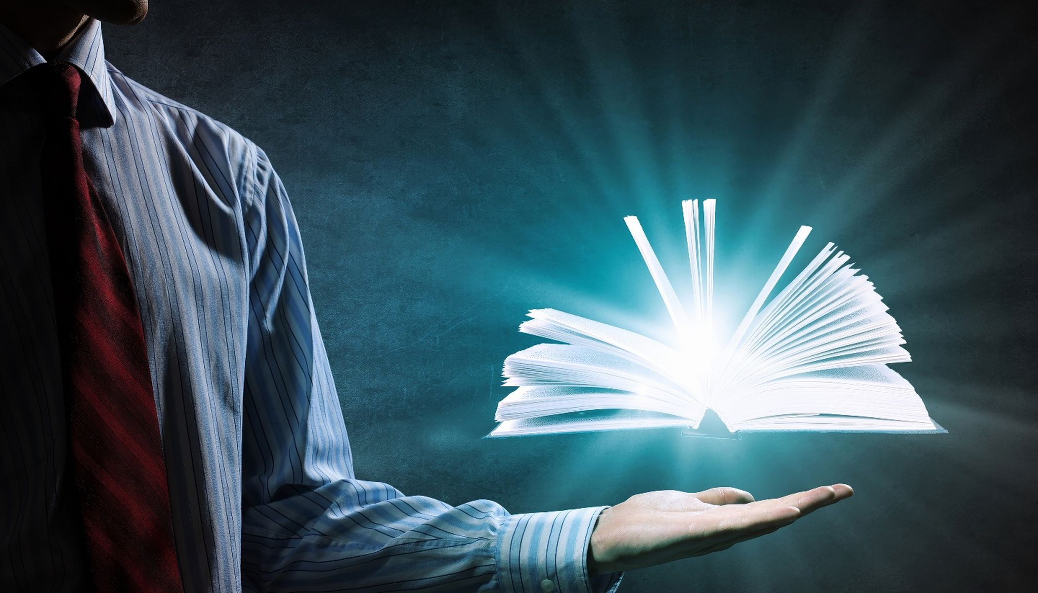 A person in a suit stretches out his hand. A book hovers above the hand, radiating bright white light and rays against a dark background.