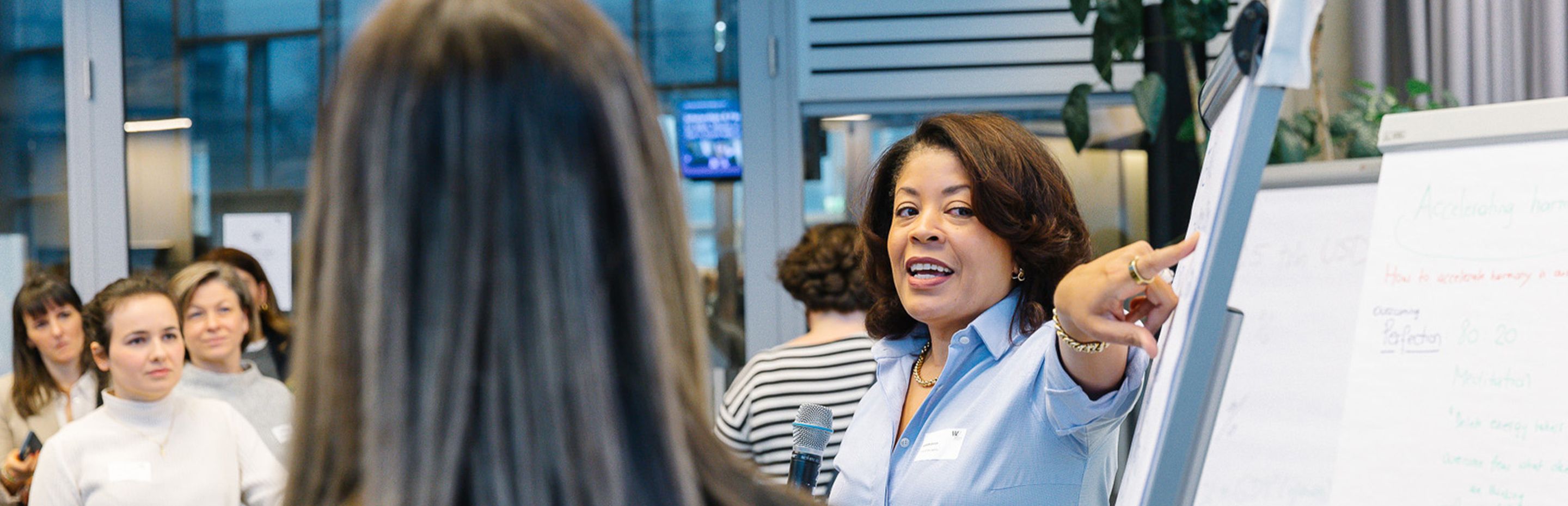 A woman in a blue shirt leads a workshop, pointing at a flip chart, with several participants listening attentively in the background. - WU Executive Academy