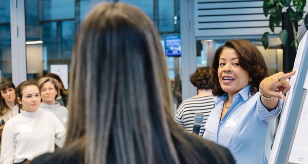 A woman in a blue shirt leads a workshop, pointing at a flip chart, with several participants listening attentively in the background. - WU Executive Academy