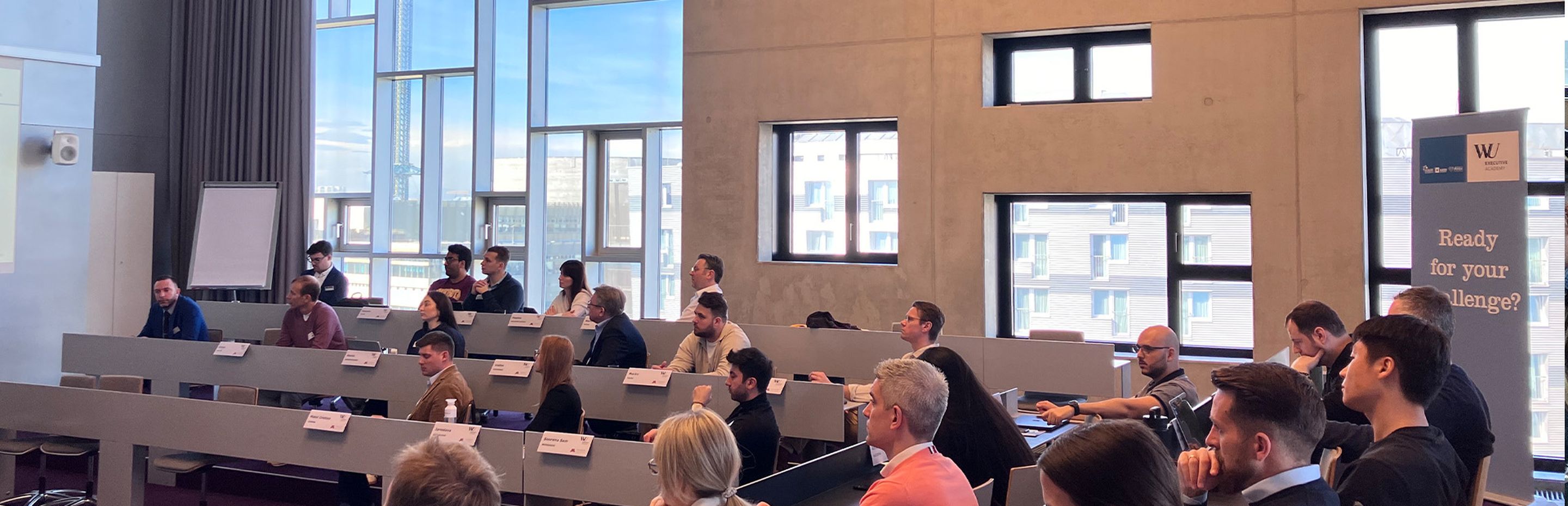 A speaker presents to an audience in a modern lecture hall with tiered seating. Attendees are engaged, with notes and laptops on desks. - WU Executive Academy