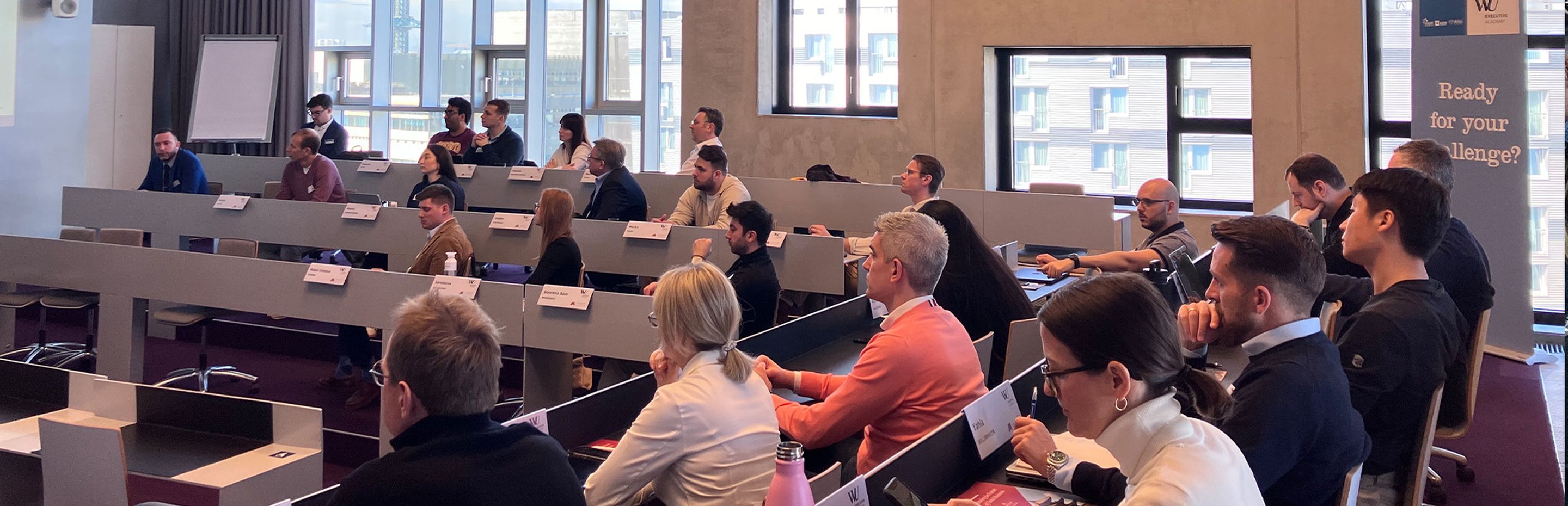 A speaker presents to an audience in a modern lecture hall with tiered seating. Attendees are engaged, with notes and laptops on desks. - WU Executive Academy